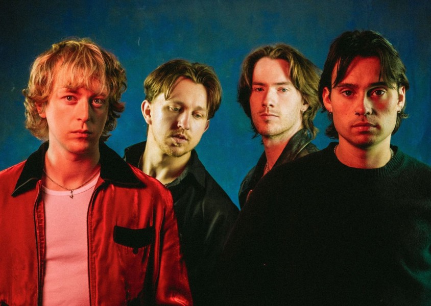 The four band members of Balancing Act stood in front of a blue background, with a red light shining towards them. Three are wearing black jackets, one is wearing a red one.x
